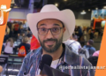 Ricardo, jornalista do Blog do Januário, veste chapéu de cowboy branco e camisa xadrez enquanto segura um microfone durante entrevista na BGS 2024. Ao fundo, o estande da Samsung no Expo Center Norte com público circulando.