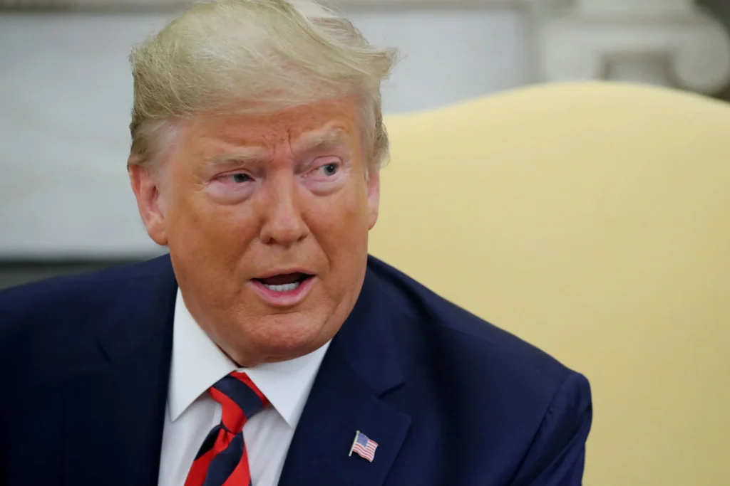Fotografia em primeiro plano de Donald Trump vestindo terno azul-marinho, camisa branca e gravata com listras vermelhas e pretas, com expressão concentrada e boca semiaberta, em ambiente interno com fundo neutro.