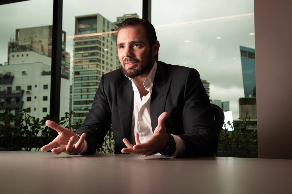 Retrato em plano médio do banqueiro Daniel Vorcaro, dono do Banco Master, vestindo terno preto e camisa branca entreaberta, sentado a uma mesa de escritório com as mãos abertas e prédios urbanos visíveis pela janela ao fundo.